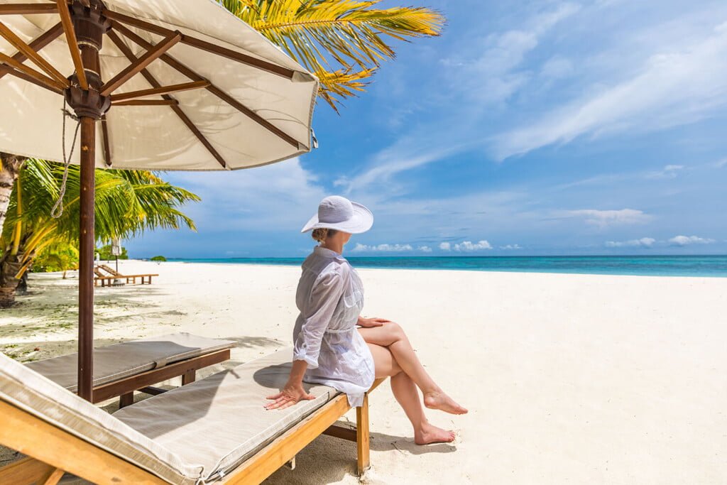 A woman lounges on a white sandy beach under a large parasol, facing the clear blue sea with a few clouds in the distance. She's wearing a white sun hat and a light, long-sleeved cover-up.